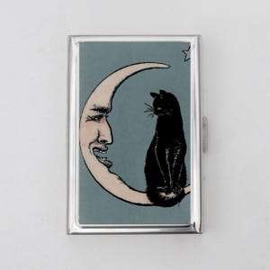 Puede incluir: Una funda de metal plateado con una ilustración vintage de un gato negro posado en una luna creciente con una cara sonriente. El fondo es de color verde azulado apagado y se ve una pequeña estrella.