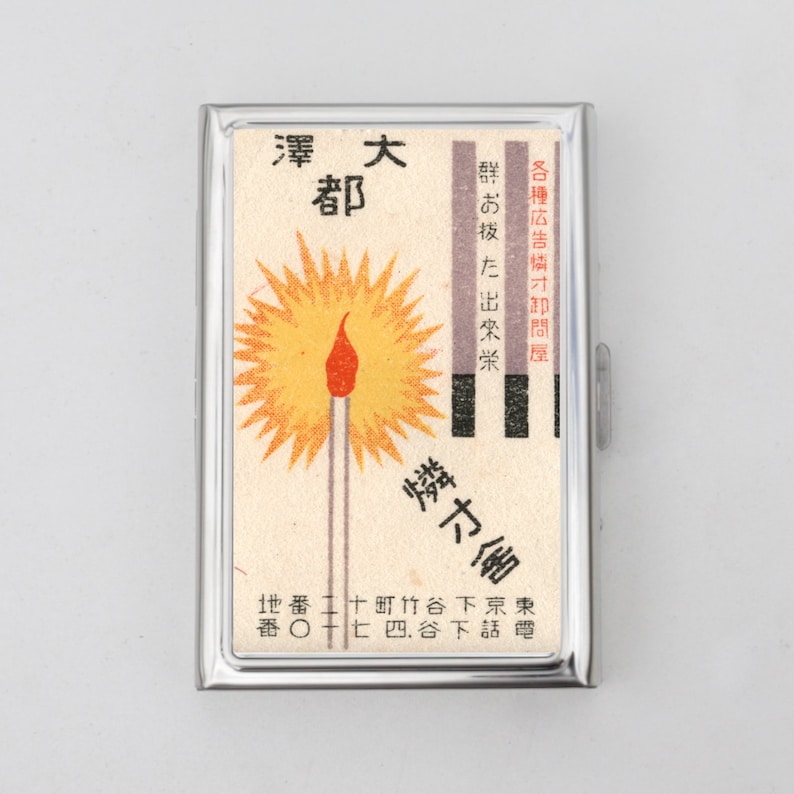 May include: A silver cigarette case with a vintage matchbox design. The case features a red and yellow illustration of a lit match with the text "澤大都" and "燐才舍" in Japanese.
