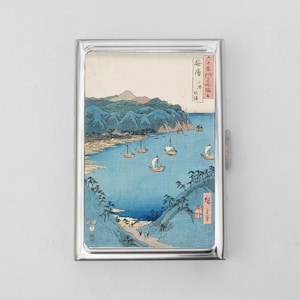 May include: A silver metal case featuring a Japanese landscape print. The artwork depicts boats sailing on a blue sea, with green hills and mountains in the background. The print has Japanese characters in the upper right corner.