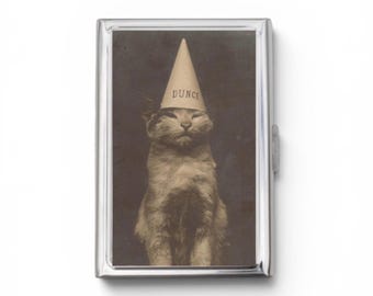 May include: A rectangular silver-toned metal case featuring a vintage-style black and white photograph of a cat wearing a conical paper hat with the word DUNCE printed in black capital letters on the front of the hat.