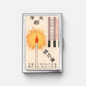 Op de afbeelding: Een rechthoekig metalen sigarettendoosje met een vintage luciferdoosje-etiket. Het etiket toont een brandende lucifer met een gele zonnestraal, Japanse tekst en een rij lucifers. Het doosje heeft een gepolijst zilverkleurig frame en een scharnierend deksel.