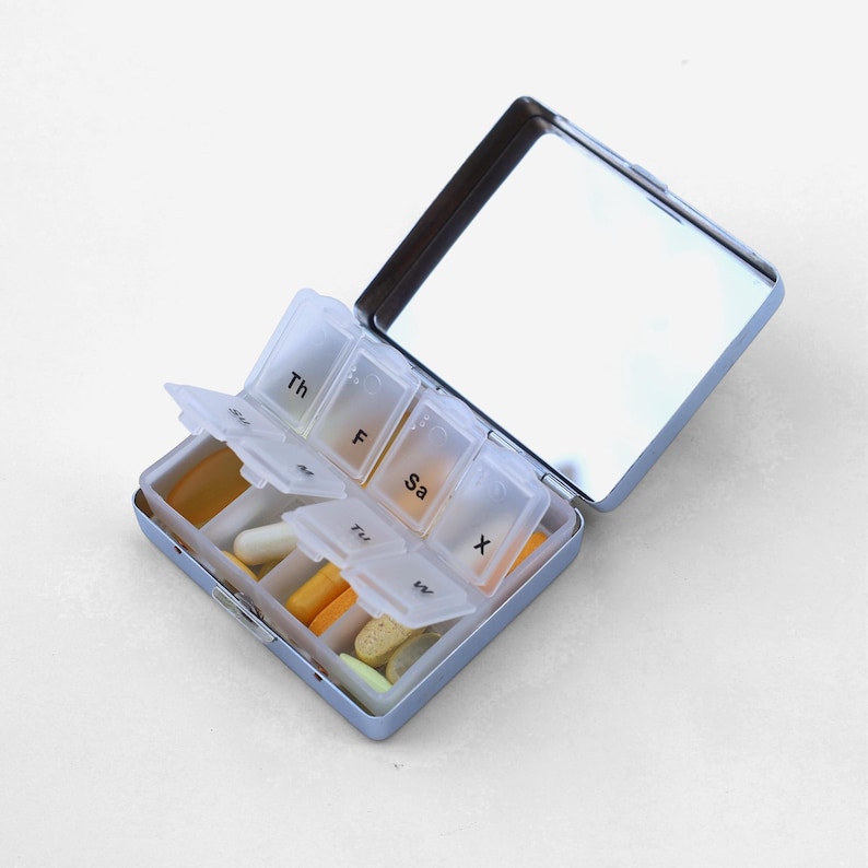 Puede incluir: Un organizador de pastillas azul con una tapa de espejo. El organizador tiene siete compartimentos etiquetados con los d&iacute;as de la semana: Th, F, Sa, Su, M, Tu, W. Los compartimentos est&aacute;n llenos de varias pastillas y c&aacute;psulas.