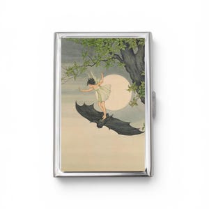 May include: A rectangular silver metal case featuring a printed illustration of a figure with wings standing on a large black bat. The scene includes a full moon, tree branches with green leaves, and a pale sky background, all encased in a polished frame.