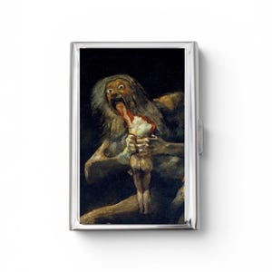 May include: A rectangular silver-toned metal case featuring a reproduction of Francisco Goya's painting Saturn Devouring His Son on the front. The artwork depicts a wide-eyed, disheveled figure consuming a smaller form against a dark background.