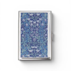 May include: A rectangular metal cigarette case featuring a polished silver frame and a decorative inlay. The inlay displays a repeating botanical pattern of blue and light purple flowers, leaves, and vines set against a deep navy blue background.