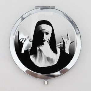 May include: A round, silver compact mirror with a black and white image of a nun making a rock and roll hand gesture.