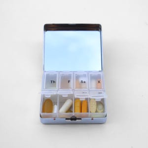 Puede incluir: Un organizador de pastillas azul con tapa de espejo. El organizador tiene siete compartimentos etiquetados con los d&iacute;as de la semana: Th, F, Sa, X. Cada compartimento contiene varias pastillas y c&aacute;psulas.