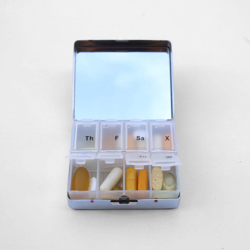 Puede incluir: Un organizador de pastillas azul con tapa de espejo. El organizador tiene siete compartimentos etiquetados con los d&iacute;as de la semana: Th, F, Sa, X. Cada compartimento contiene varias pastillas y c&aacute;psulas.