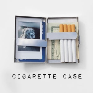 May include: A rectangular silver metal cigarette case with a hinged opening. The left side holds a square photograph of a dog and a blue credit card. The right side contains four cigarettes and a folded one hundred dollar bill held by a metal clip.