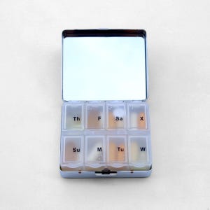 Puede incluir: Una caja de pastillas de plata con un espejo en la parte superior. La caja de pastillas tiene siete compartimentos etiquetados con los d&iacute;as de la semana: Su, M, Tu, W, Th, F, Sa.