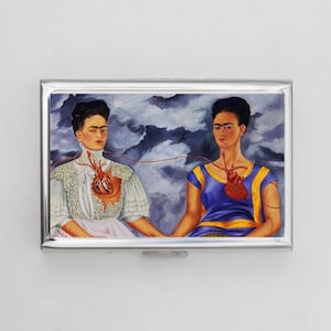 May include: A rectangular, silver-framed case with a painting of two women. Each has an exposed heart, connected by a red string. One wears a white dress, the other a blue top. The background shows a cloudy sky.
