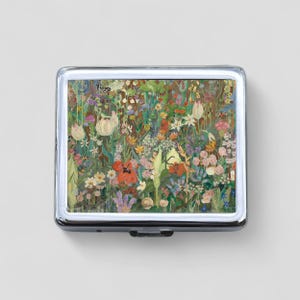 May include: A silver compact with a colorful floral design featuring a variety of flowers, butterflies, and leaves. The compact has a hinged lid and a silver clasp.