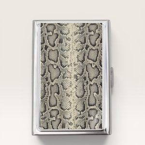 May include: A rectangular metal cigarette case featuring a polished silver-tone frame and a central inlay panel with a detailed black, gray, and cream-colored snakeskin pattern. The case has a hinged design with a small side latch for closure.