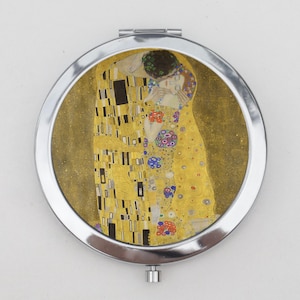 May include: A silver compact mirror with a round, gold-toned image of Gustav Klimt's "The Kiss" painting. The painting depicts a couple embracing in a golden, mosaic-like pattern.