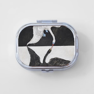 May include: A silver compact mirror with a black and white image of two swans, one white and one black, facing each other.