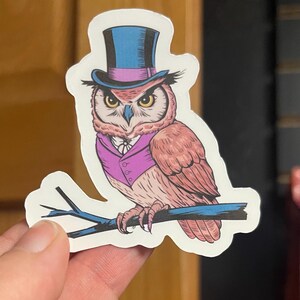 May include: A sticker of a cartoon owl wearing a top hat and a purple vest. The owl is perched on a branch with its wings spread out.