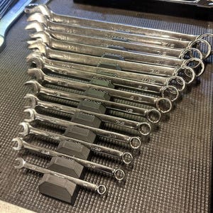 May include: A set of silver wrenches arranged in a row. Each wrench has a different size, with measurements like 7/8 and 1/2. The wrenches are neatly organized on a dark surface, suggesting a tool set for mechanical work.