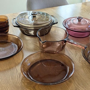 May include: A collection of brown glass cookware, including a casserole dish, a frying pan, a pie plate, and a saucepan. The cookware is arranged on a wooden table.