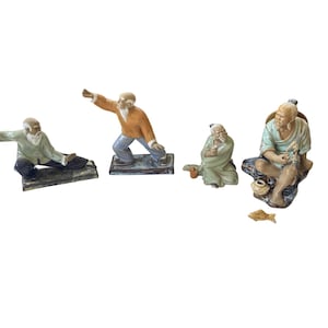 May include: A set of four ceramic figurines depicting elderly figures in various poses. Two figures are in active stances, one is seated holding a cup, and one is seated fishing with a small basket and a separate fish figurine placed nearby.