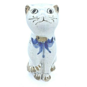 May include: A white ceramic cat figurine with blue and gold painted details. The cat is sitting with its tail curled around its legs. It has a blue and gold bow around its neck.