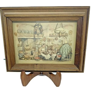 May include: A framed print depicting a kitchen scene with two women, one seated and the other standing. The print is in a wooden frame and is displayed on a wooden easel. The kitchen features a fireplace, hanging pots, and various kitchenware.