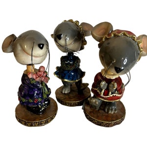 May include: Three gray ceramic mouse figurines wearing dresses. The mouse on the left is wearing a purple dress with pink flowers, the middle mouse is wearing a blue dress with green trim, and the mouse on the right is wearing a red dress with white trim.