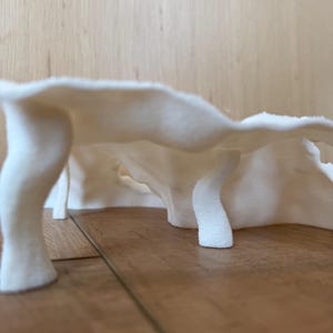 May include: A low-angle shot of a white, abstract sculpture with a textured surface. The sculpture has a curved, organic shape supported by several cylindrical legs. The background is a light-colored wooden surface.