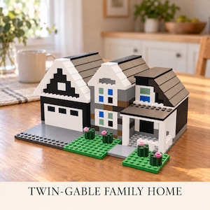 May include: A building block model of a house titled TWIN-GABLE FAMILY HOME. The structure features a black and white garage, a grey multi-storey section with blue and green windows, a covered porch, and green base plates with small pink flower accents.