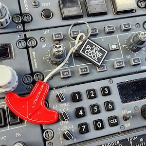 Könnte beinhalten: Nahaufnahme eines Flugzeugcockpit-Bedienfelds mit verschiedenen Knöpfen, Schaltern und Zifferblättern. Ein roter Griff und ein Schlüsselanhänger mit dem Text "JUST PLANE COOL" sind sichtbar. Das Panel ist grau mit schwarzen und weißen Details.