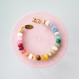 Puede incluir: Una pulsera de cuentas coloridas con un cierre y detalles dorados. La pulsera presenta una variedad de cuentas de piedras preciosas en un arcoíris de colores, incluyendo rosa, rojo, naranja, amarillo, verde, azul y blanco. La pulsera se muestra sobre una superficie circular rosa.