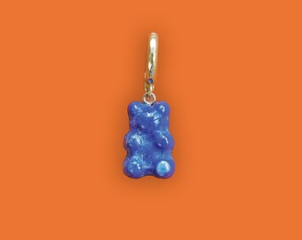 May include: A single blue gummy bear charm earring with a gold-coloured hoop and a small blue gem. The earring is set against a bright orange background.