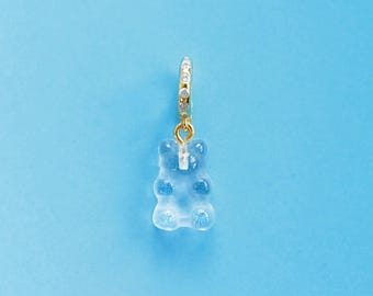 May include: A gold-coloured hoop earring featuring a clear, gummy bear charm. The charm is light blue and shaped like a bear. The earring is set against a solid light blue backdrop.