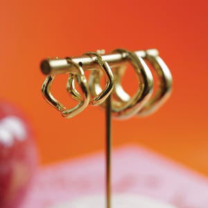 May include: A selection of gold-coloured hoop earrings displayed on a gold-coloured jewellery stand. The earrings have a distinctive, square-like form. The background is a gradient of orange and pink.