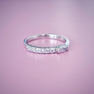 May include: A silver ring with a row of small, round, clear stones and a single, rectangular, clear stone. The ring is set against a soft, pink background.