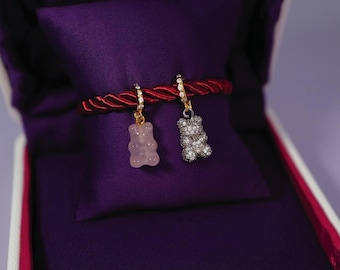 May include: Two gold hoop earrings, each with a gummy bear charm. One charm is a pale pink, translucent stone, the other is covered in small, sparkling crystals. The earrings are presented in a purple jewellery box.