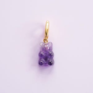 May include: A gold-coloured hoop earring featuring a purple gummy bear charm. The translucent purple gummy bear has darker purple detailing. The earring is set against a light purple backdrop.