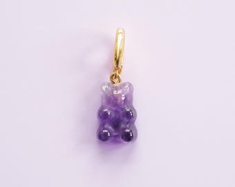 May include: A gold-coloured hoop earring featuring a purple gummy bear charm. The translucent purple gummy bear has darker purple detailing. The earring is set against a light purple backdrop.