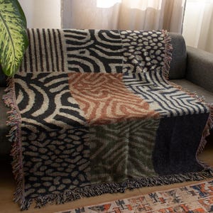 Könnte beinhalten: Eine Patchwork-Decke mit verschiedenen geometrischen und Tiermustern in Schwarz-, Braun- und Beigetönen. Die Decke ist über ein graues Sofa drapiert und hat einen Fransenrand. Ein dekoratives Heimtextil.