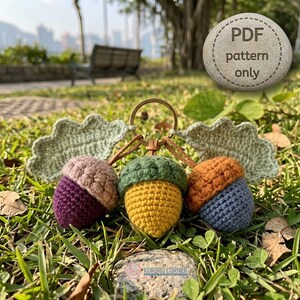 Puede incluir: Un llavero de ganchillo con tres bellotas con sombreros texturizados y cuerpos coloridos en morado, amarillo y azul, unidos a una anilla de metal con dos hojas de ganchillo verde claro. Una piedra muestra el texto PDF pattern only.