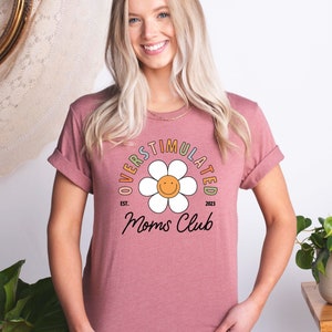 May include: A pink t-shirt with a white daisy and the text "Overstimulated Moms Club Est. 2023" in rainbow colors.