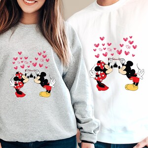 May include: Two sweatshirts, one gray and one white, feature a cartoon design of Mickey and Minnie Mouse kissing, with pink hearts floating above them. The design includes the word "Believe" and a castle silhouette.