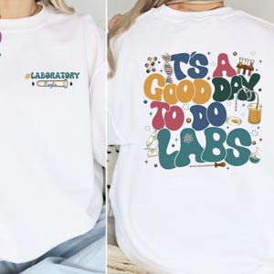 May include: White sweatshirt with the text "#LABORATORY Kayla" on the front and "IT'S A GOOD DAY TO DO LABS" on the back. The design includes lab equipment illustrations in various colors.