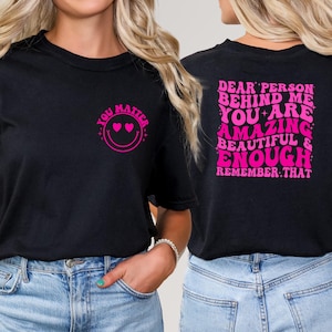 May include: Two black t-shirts with pink text. The front shirt says "You Matter" with a smiley face and hearts for eyes. The back shirt says "Dear Person Behind Me You Are Amazing Beautiful & Enough Remember That".