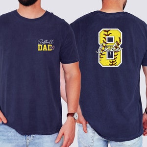 May include: A navy blue t-shirt with the text "Softball Dad" on the front and a yellow softball with the number 8 and the text "Dexter" on the back.