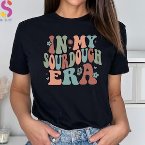 May include: Black t-shirt with the text "IN MY SOURDOUGH ERA" in retro-style lettering. The design includes floral and butterfly accents in shades of green, orange, and blue. The shirt is a casual wear item.