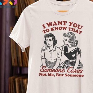 Op de afbeelding: Wit T-shirt met een zwart-witte illustratie van twee vrouwen. De tekst op het shirt luidt "Ik wil dat je weet dat iemand geeft, niet ik, maar iemand."
