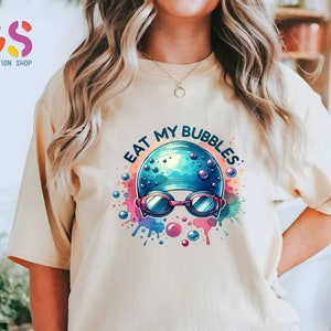 May include: A light beige t-shirt with a colorful graphic of a swimmer wearing a blue swim cap and goggles with the text "Eat My Bubbles".