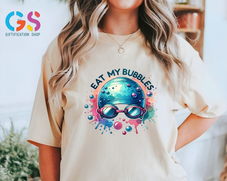 May include: A light beige t-shirt with a colorful graphic of a swimmer wearing a blue swim cap and goggles with the text "Eat My Bubbles".