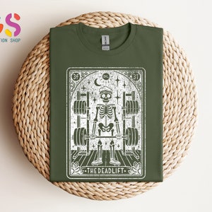 May include: A green t-shirt featuring a white graphic of a skeleton lifting weights. The graphic is in the style of a tarot card and reads "The Deadlift" at the bottom.