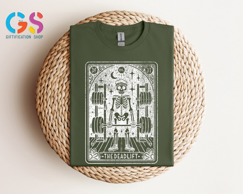 May include: A green t-shirt featuring a white graphic of a skeleton lifting weights. The graphic is in the style of a tarot card and reads "The Deadlift" at the bottom.